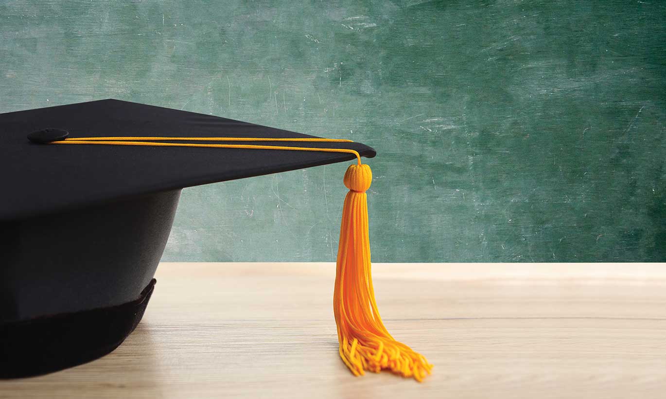 A black graduation cap with a gold tassell sits in front of a chalkboard