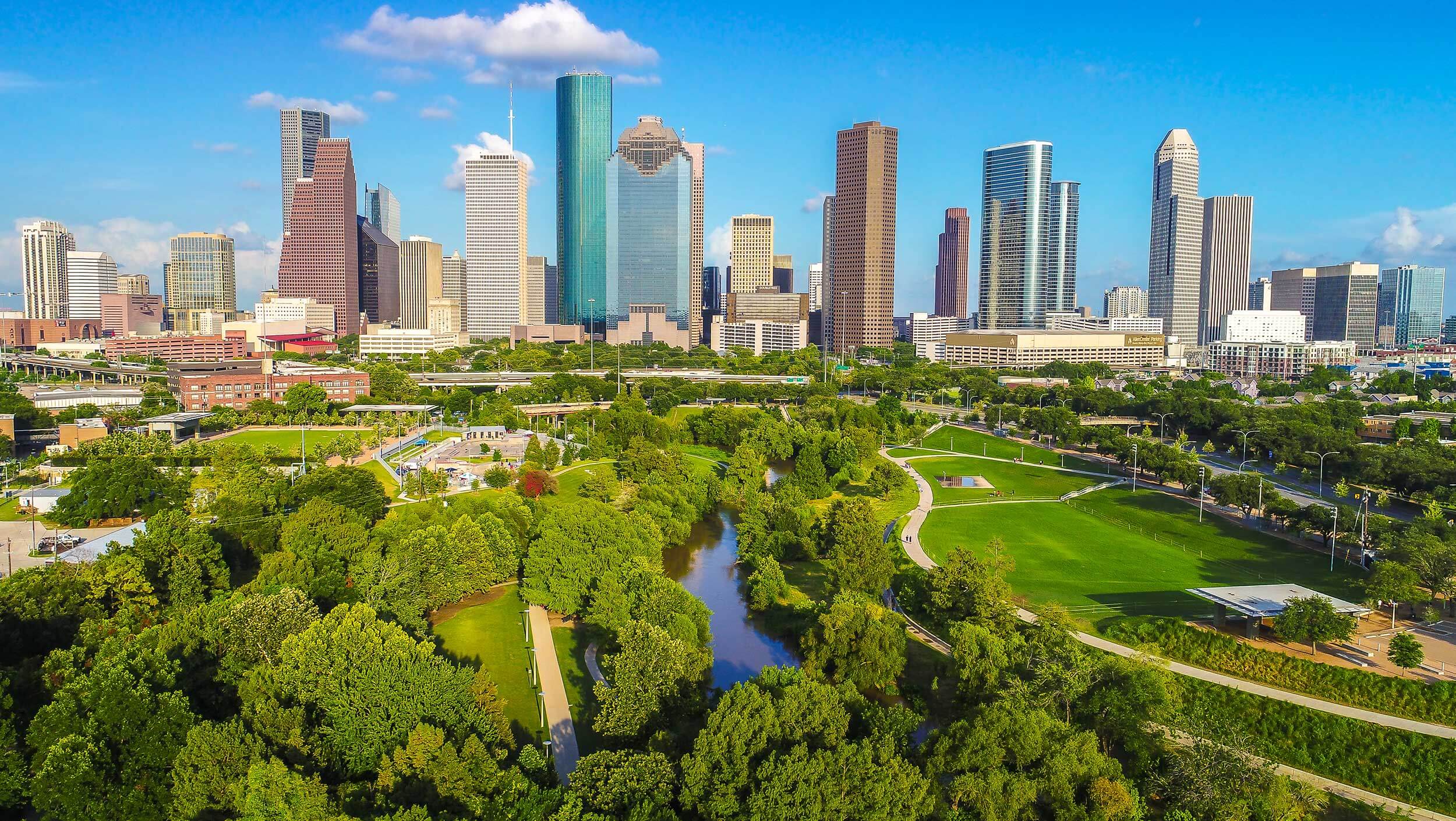 Beautiful green spaces and a river run adjacent to the tall buildings in downtown Houston, Texas