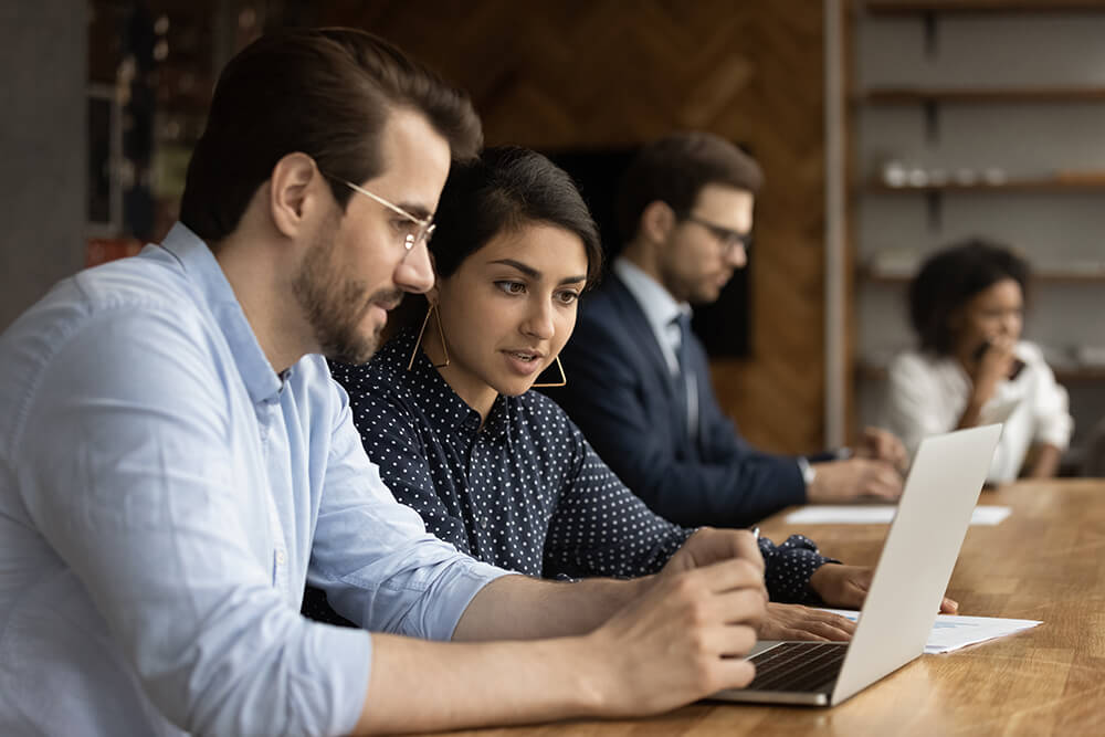 Focused male millennial intern look at laptop screen as he listens to skilled Indian female mentor